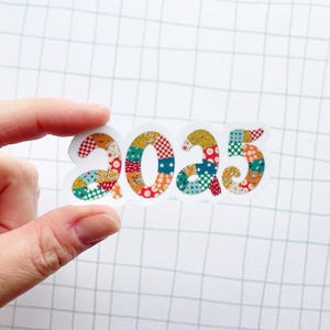 May include: A close-up of a hand holding a sticker with the year 2025 written in colorful patchwork fabric.