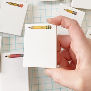 May include: A set of small white notepads with a pencil illustration on each. The pencils are yellow, red, and brown with a black eraser.