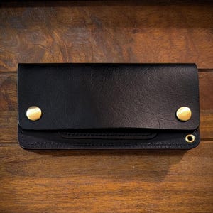 May include: Black leather wallet with a snap closure and two gold-coloured rivets. The wallet is rectangular and has a flap that folds over the main compartment. The background is a wooden surface.