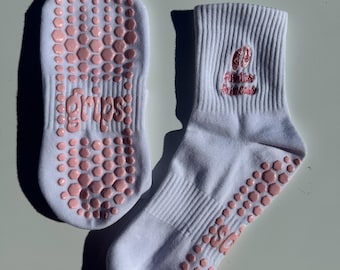 May include: A pair of white ankle socks with pink non-slip grips on the bottom. The socks have a pink logo that says "Fitness Addict" on the top of one sock and "Grips" on the bottom of the other sock.