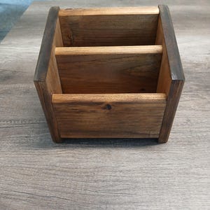 May include: A dark brown stained wooden desk organizer featuring two distinct compartments with a taller back section and a shorter front section, constructed with visible wood grain and a rectangular base with four small feet.