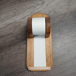 May include: A wooden paper roll dispenser featuring a rectangular base with rounded ends, two upright supports holding a white paper roll, and a light-colored wooden strip secured at the bottom with two small metal fasteners to hold the paper flat.