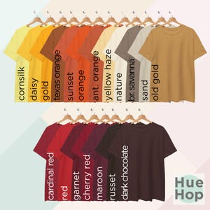 May include: A collection of 20 t-shirts in various shades of orange, red, and brown, hanging on hangers. The t-shirts are labeled with color names like "cornsilk", "daisy", "gold", "texas orange", "sunset", "orange", "ant. orange", "yellow haze", "nature", "br. savanna", "sand", "old gold", "cardinal red", "red", "garnet", "cherry red", "maroon", "russet", "dark chocolate", and "Hue Hop".