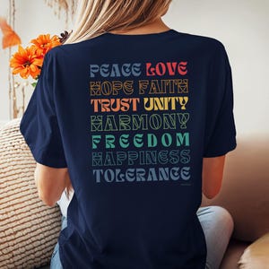 May include: A navy blue t-shirt with a rainbow-colored graphic on the back. The graphic features the words "Peace Love Hope Faith Trust Unity Harmony Freedom Happiness Tolerance".