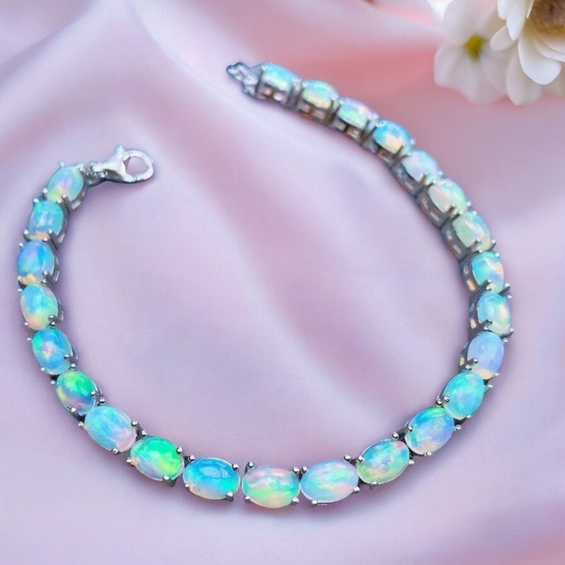 Op de afbeelding: Een zilveren armband met een reeks ovale opalen. De opalen tonen blauwe, groene en witte tinten, gezet in een zilveren ketting. De armband wordt gepresenteerd op een roze satijnen oppervlak.