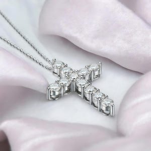 May include: A silver-toned cross pendant necklace with a delicate chain. The cross is adorned with multiple clear, round-cut gemstones, creating a sparkling effect. The necklace rests on a white, silky fabric.