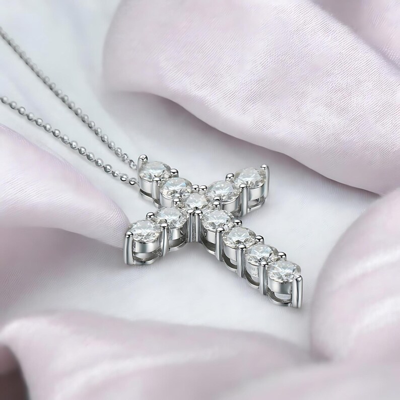 May include: A silver-toned cross pendant necklace with a delicate chain. The cross is adorned with multiple clear, round-cut gemstones, creating a sparkling effect. The necklace rests on a white, silky fabric.