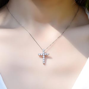 May include: A silver necklace with a cross pendant. The cross is encrusted with small, clear gemstones. The necklace is delicate and appears to be made of silver. The pendant is centered on the chest.