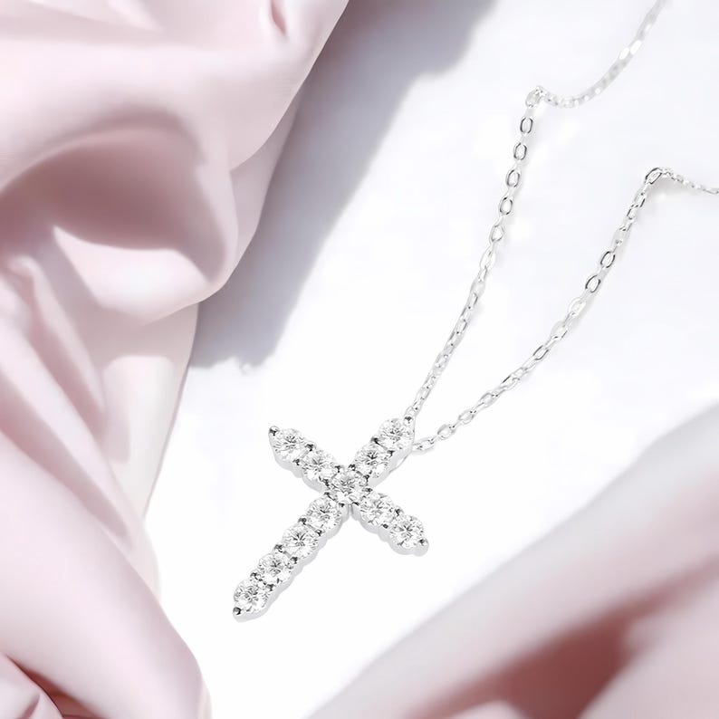 May include: A silver cross pendant necklace with a delicate chain. The cross is encrusted with sparkling, clear gemstones. The necklace is displayed on a white surface with a soft pink fabric in the background, creating a luxurious feel.