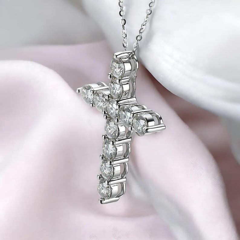 May include: A silver cross pendant necklace with clear, round-cut gemstones. The cross is made of silver and features multiple gemstones set in a cross shape. The necklace is displayed on a soft, light pink and white background.