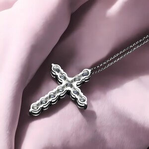 May include: A silver cross pendant necklace with a delicate chain. The cross is encrusted with multiple clear gemstones. The necklace is displayed on a pink fabric background.