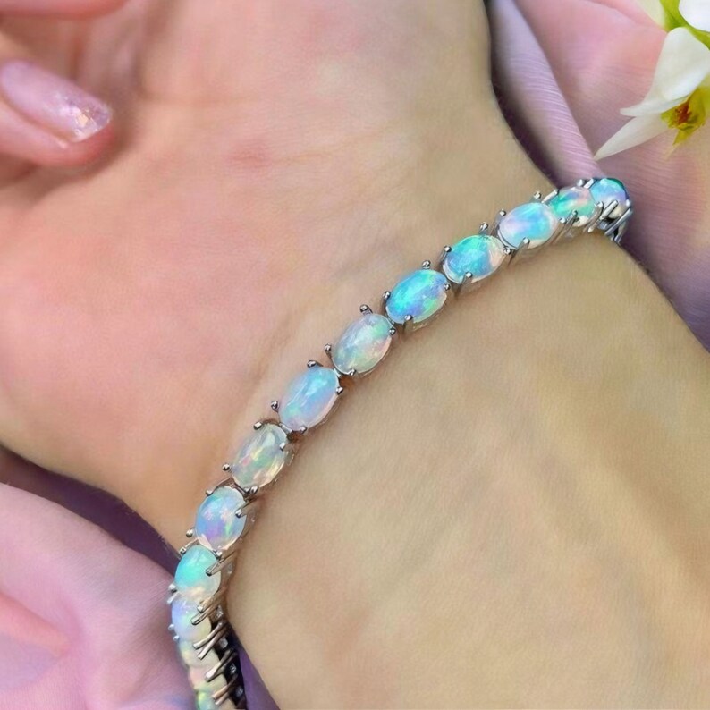 Op de afbeelding: Een zilveren armband met een reeks ovale opalen. De opalen vertonen een kleurenspel met flitsen van blauw, groen en geel. De armband is gezet op een zilveren ketting, waardoor een delicaat en elegant sieraad ontstaat.