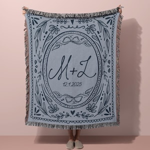 Può includere: Una coperta tessuta blu chiaro con un bordo decorativo e frange. Il centro presenta un design ovale con le iniziali "M + L" e la data "12.1.2025" in scrittura blu scuro. Motivi floreali e di foglie circondano il design centrale.