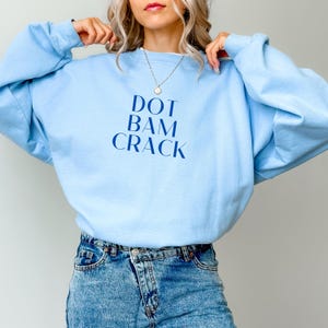 Puede incluir: Sudadera azul claro con el texto "DOT BAM CRACK" en azul oscuro. Se combina con vaqueros azules. El estilo general es informal y cómodo.
