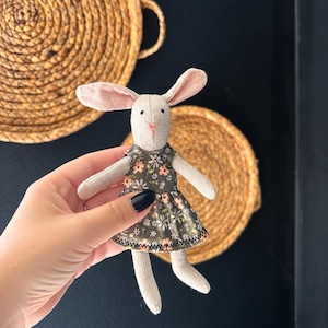 May include: A small stuffed rabbit toy made of light beige fabric with pink inner ears and a stitched pink nose. The rabbit is wearing a dark olive green dress featuring a repeating pattern of small white and peach-colored flowers.