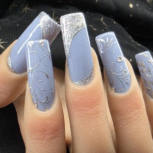 Puede incluir: Primer plano de cinco uñas largas, de color azul claro, con puntas de brillo plateado y diseños florales plateados.