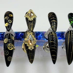 May include: Seven black stiletto nails with gold accents. The nails are decorated with gold glitter, gold gears, and a gold diamond-shaped rhinestone.