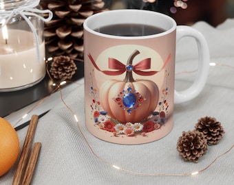May include: White ceramic mug featuring a pumpkin design with a blue jewel, red bow, and floral accents. The mug has a white handle and is filled with a dark liquid. The design is set against a light pink background.