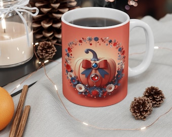 May include: A white ceramic mug with a pumpkin design. The pumpkin is decorated with a blue bow and surrounded by a floral wreath in red, white, and blue. The mug is filled with a dark liquid.