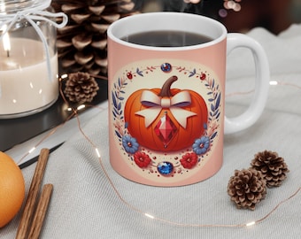 May include: White ceramic mug featuring an orange pumpkin design with a white bow and a red gem. The pumpkin is surrounded by a floral wreath with blue and red accents. The mug has a pink background and is filled with a dark liquid.