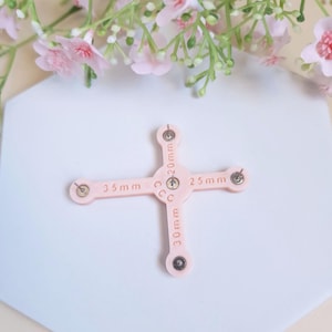 May include: A pink plastic ring sizer with four arms, each marked with a different size in millimeters: 25mm, 30mm, 35mm, and 40mm. The sizer is on a white hexagonal surface.