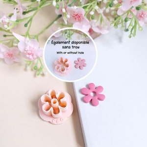 May include: Two pink flower-shaped cookie cutters, one with a hole in the centre and one without. The text "Également disponible sans trou" and "With or without hole" is visible in the image.