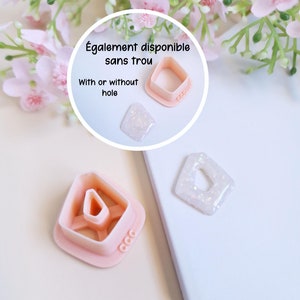 Puede incluir: Dos cortadores de galletas de plástico rosa en forma de diamante, uno con un agujero en el centro y otro sin. Los cortadores están sobre una superficie blanca con un fondo rosa. El texto "Également disponible sans trou" es visible en la imagen. El texto "With or without hole" también es visible en la imagen.
