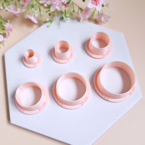 Peut inclure: Ensemble de six emporte-pièces ronds en plastique rose sur une surface hexagonale blanche. Les emporte-pièces sont de tailles différentes, le plus grand mesurant 4,5 cm de diamètre et le plus petit 1,5 cm de diamètre.