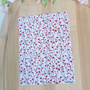 Peut inclure: Une carte rectangulaire avec un fond blanc, ornée d'un motif de fleurs rouges en forme de cœur sur de fines tiges noires. La carte porte le texte "CamClayCo" dans le coin inférieur droit. Des fleurs roses et de la verdure sont en arrière-plan.