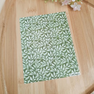 Puede incluir: Un papel rectangular con un diseño de hojas verdes y blancas. Está colocado sobre una tabla de madera, con flores rosas y blancas en el fondo. El texto "CamClayCo" se encuentra en la esquina inferior derecha.