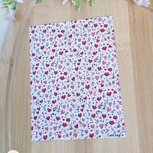 Peut inclure: Une carte blanche ornée d'un motif répété de cœurs et de fleurs rouges. Le motif comprend de petits cœurs rouges et de plus grandes fleurs en forme de cœur avec des tiges et des feuilles noires. La carte est posée sur une surface en bois, avec des fleurs roses en arrière-plan. Le texte "CamClayCo" est visible.