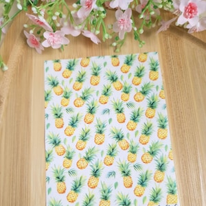 Puede incluir: Tela blanca con un patrón repetido de piñas amarillas y hojas verdes. La tela tiene el texto "CamClayCo" en la esquina inferior derecha.