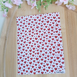 Peut inclure: Une feuille rectangulaire avec un motif de cœurs rouges sur fond blanc. Les cœurs sont de tailles variées et espacés uniformément. La feuille est posée sur une surface en bois, avec des fleurs roses en arrière-plan. Le texte "CamClayCo" est visible.