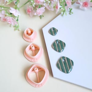 May include: A selection of pink polymer clay cutters and green and gold striped earrings. The cutters are square-shaped with a central peg and three small circles on the side. The earrings have a rounded square shape.