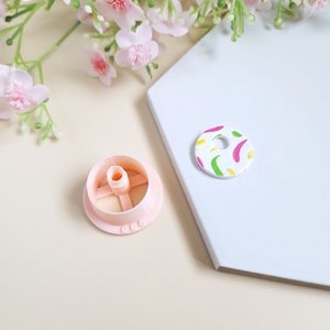 May include: A pink plastic cake decorating tool with a round base and a centre post. The tool is designed to create a hole in the centre of a cake. A round, white cookie with a colourful abstract design is also shown.