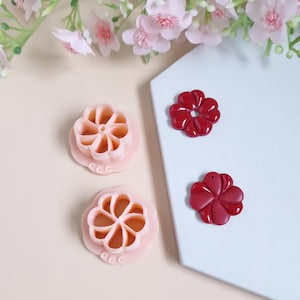 May include: Two pink plastic flower-shaped cookie cutters and two red polymer clay flower charms. The cookie cutters have a raised design and the charms have a smooth finish.