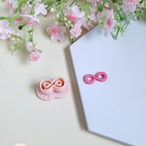 May include: A pink infinity symbol cookie cutter and a separate infinity symbol. The cookie cutter has a circular base with three small circles. Pink flowers and greenery are in the background. The objects are on a white and beige surface.