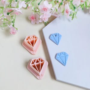 Puede incluir: Dos cortadores de galletas rosas en forma de diamante y dos adornos azules claros en forma de diamante se muestran sobre una superficie blanca. Los cortadores tienen un diseño interior en forma de diamante. Flores rosas en el fondo.