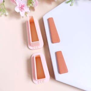 Peut inclure: Deux moules en plastique rose pour faire des formes en pâte à sucre ou en argile polymère. Les moules sont en forme de rectangle aux coins arrondis. Les moules ont un motif en relief sur le fond. Il y a deux formes en argile brune sur une surface blanche.