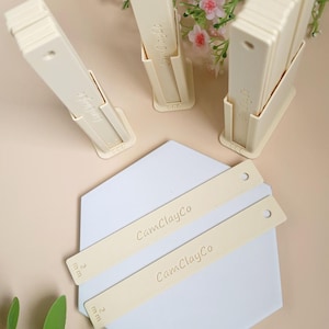 Könnte beinhalten: Zwei Sätze weißer Kunststoffwerkzeuge für Polymer Clay mit einem Lineal, das in Millimetern gekennzeichnet ist. Die Werkzeuge sind mit "CamClayCo" beschriftet und auf einer weißen sechseckigen Oberfläche angeordnet.
