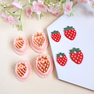 Könnte beinhalten: Vier rosa Plastik-Ausstecher in Form von Erdbeeren und zwei rote und weiße Erdbeer-Ohrringe aus Polymer Clay.
