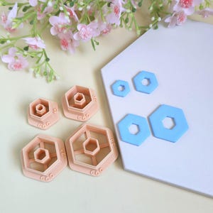 May include: A set of four peach-coloured hexagonal clay cutters with varying sizes and four corresponding light blue hexagonal clay shapes. The cutters feature embossed numerical size markings and the brand name CamClayCo on the base.