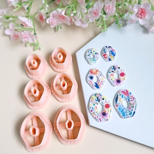 Puede incluir: Un juego de cinco cortadores de galletas de plástico rosa con un diseño floral y el texto "CCC" en ellos. También hay seis pendientes de arcilla polimérica con estampado floral de colores sobre una superficie blanca.