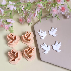 May include: A selection of dove-shaped cookie cutters and white dove embellishments are arranged on a surface. The cutters are a peach hue, and the embellishments are white. Pink flowers are in the background. The logo "CamClayCo" is visible.