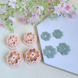 Peut inclure: Ensemble de découpoirs à biscuits en forme de fleurs roses et de fleurs découpées vertes. Les découpoirs présentent un motif de pétales détaillé et le texte "CCC" sur la base. Des fleurs roses sont en arrière-plan.