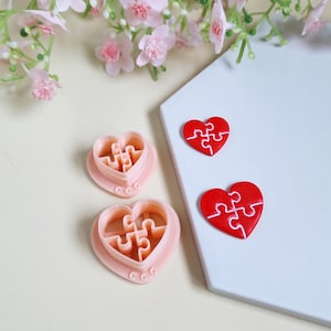 Peut inclure: Emporte-pièces roses en forme de cœur et de puzzle, ainsi que deux pièces de puzzle rouges en forme de cœur. Les pièces du puzzle sont sur une surface blanche, et les emporte-pièces sur une surface beige clair. Fleurs roses en arrière-plan.