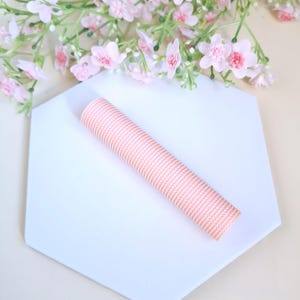 May include: A light pink, textured, cylindrical object rests on a white, hexagonal surface. Delicate pink flowers with green stems and leaves are in the background. The object appears to be a decorative item or craft supply.