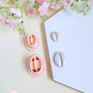 Può includere: Due formine per biscotti rosa chiaro, a forma ovale, con disegni interni e due decorazioni argentate a forma di conchiglia. Gli oggetti sono disposti su una superficie bianca, con fiori rosa sullo sfondo. Le formine hanno un bordo smerlato.