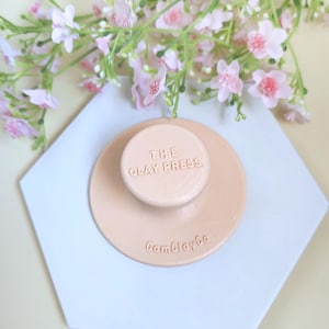Könnte beinhalten: Hellpfirsichfarbene Tonpresse mit dem Text "THE CLAY PRESS" und "CamClayCo" geprägt. Sie liegt auf einer weißen sechseckigen Fliese, mit rosa Blüten im Hintergrund. Für Bastelarbeiten.