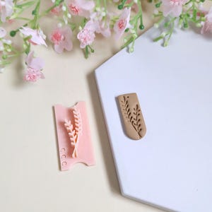 Peut inclure: Un tampon en argile polymère rose avec un motif de blé et un pendentif en argile polymère marron avec un motif de blé. Le tampon est rectangulaire avec un motif de blé et trois petits cercles. Le pendentif est un rectangle arrondi.